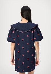Femme portant une robe bleu marine avec des motifs de cœurs rouges, des manches bouffantes et un grand col froncé, vue de dos.