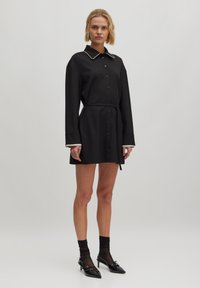 Schwarzes Hemdkleid mit langen Ärmeln, weißen Paspeln am Kragen und an den Manschetten, tailliert, knielang, aus einem glatten Stoff gefertigt.