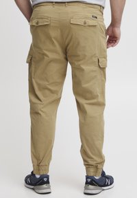Khaki cargo byxor med elastiska muddar, har flera fickor och en texturerad yta. Bärs med mörka sneakers, sedd bakifrån.