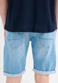 Shorts de mezclilla de un azul claro con dobladillos enrollados, que cuentan con dos bolsillos en la parte posterior y una textura suave. Se llevan con una camisa oscura texturizada.