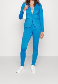 Modrý blazer a odpovídající kalhoty z hladkého materiálu. Má slim fit střih s dlouhými rukávy, v kombinaci s bílými teniskami a malou kabelkou.