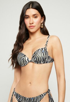 Frau trägt einen schwarz-weiß gestreiften Zebra-Bikini, goldene Creolen, lange dunkle Haare, vor einfachem hellem Hintergrund.