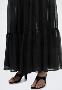 Μαύρη διαφανής μάξι φούστα με μαζεμένες στρώσεις, φορεμένη με μαύρα πέδιλα με τακούνι ανοιχτά μπροστά και σχεδιασμό με λουράκι μεταξύ δαχτύλων.