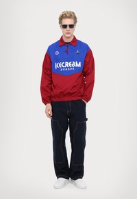 Giacca a vento rossa e blu con colletto e vita elasticizzata, con logo "ICECREAM EUROPE". Abbinata a pantaloni scuri e sneaker bianche.
