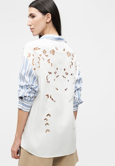 Γυναίκα φόρεσε λευκό πουκάμισο με μπλε ριγέ μανίκια και κεντήματα με λουλούδια στην πλάτη, συνδυασμένο με μπεζ παντελόνι.