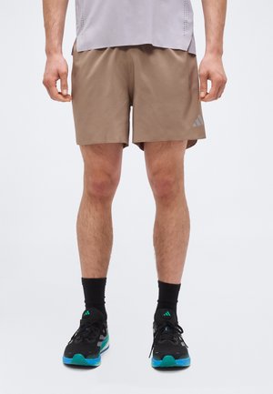 Man draagt lichtbruine sportshorts, zwarte sokken tot halverwege de kuit en zwarte hardloopschoenen met turquoise en blauwe accenten.