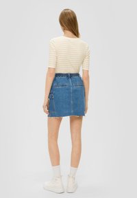 Denimrock mit fransigem Saum, zwei hinteren Taschen und einer Seiten Tasche. Kombiniert mit einem cremefarben gestreiften Kurzarmoberteil und weißen Turnschuhen.