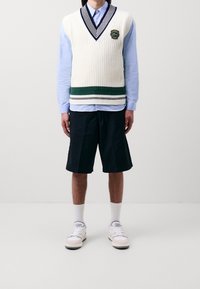 Kremowa dzianinowa kamizelka z dekoltem w serek w granatowe paski, zielony poziomy pas; jasnoniebieska koszula na guziki, czarne szorty, białe trampki.