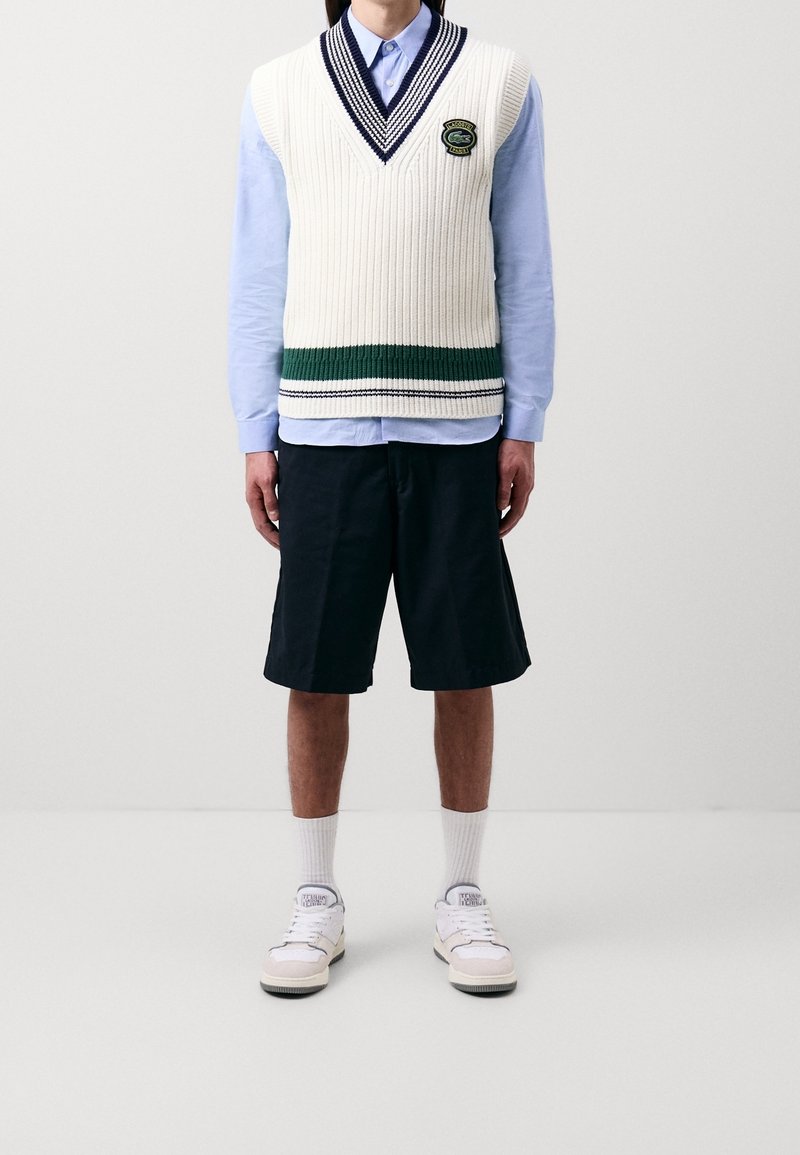 Kremowa dzianinowa kamizelka z dekoltem w serek w granatowe paski, zielony poziomy pas; jasnoniebieska koszula na guziki, czarne szorty, białe trampki.