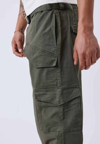 Pantaloni cargo verde oliva realizzati in tessuto resistente, dotati di molteplici tasche laterali e un discreto dettaglio del logo. I passanti per la cintura sono visibili in vita.