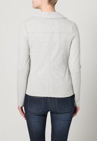 Jasnoszara kurtka z długim rękawem i kołnierzem, o gładkiej fakturze i detalach szwu z tyłu. W połączeniu z ciemnymi jeansami.