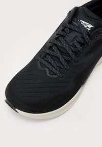 Black athletic shoe featuring a mesh upper, textured design, contrasting white sole, and flat black laces with subtle reflective accents.
