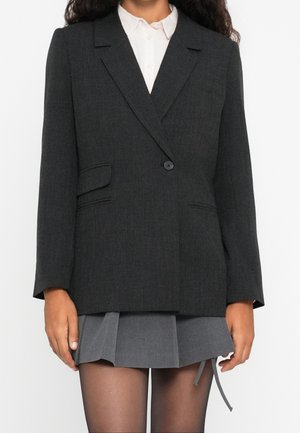 Kobieta ubrana w ciemnoszary blezer na białej koszuli i krótką plisowaną szarą spódnicę z czarnymi przezroczystymi rajstopami, stojąca na białym tle.