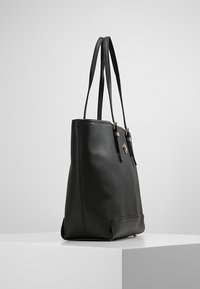 Černá kožená taška přes rameno s dvojitými ramenními popruhy a kovovým logem, vystavená na bílém podstavci na jednobarevném pozadí.