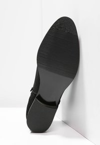 Suela de zapato negra con agarre texturizado, posicionada en ángulo sobre un fondo blanco, mostrando su diseño y detalles de material.
