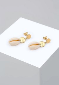 Boucles d'oreilles clous en or avec des disques ronds martelés et des breloques coquillages blanc cassé attachées, présentées sur un socle blanc sur un fond gris clair.