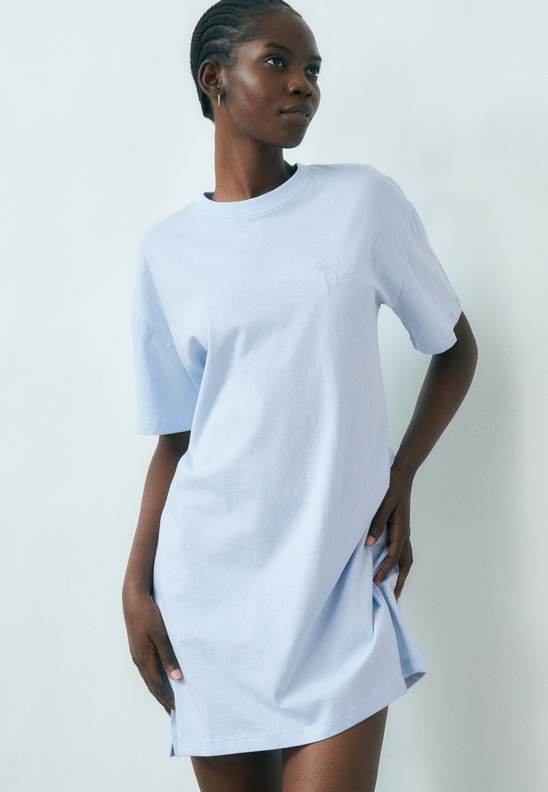 Γυναίκα με πλεξούδες, φοράει ανοιχτό μπλε, φαρδύ φόρεμα τύπου μπλούζας, στέκεται μπροστά σε απλό ανοιχτό φόντο, με το ένα χέρι στη μέση.