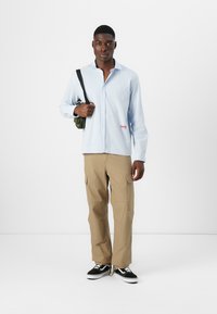Light blue checkered button-up shirt, tan cargo pants, and black sneakers with white soles. Model carries a small green shoulder bag.