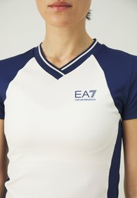 Športna kratka majica s kratkimi rokavi v beli in mornarsko modri barvi z obliko v izrezu v obliki črke V. Vsebuje logotip EA7 na levi strani prsnega koša in kontrastne šivane detajle.