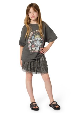 Niña con cabello largo que lleva una camiseta gráfica oscura y de gran tamaño, falda plisada brillante y sandalias negras, de pie con una mano en la cadera.