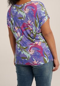 Krátký rukáv top v purpurové látce s květinovým vzorem v růžových, zelených a modrých akcenty, kombinovaný s modrými džíny.