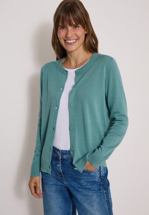 Femme souriante portant un cardigan bleu sarcelle boutonné sur un haut blanc et un jean bleu, debout avec une main dans la poche sur un fond clair.