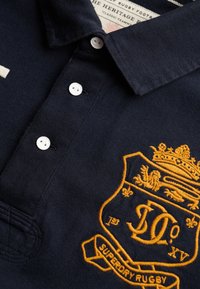 Polo bleu marine avec des boutons blancs et un blason brodé en or représentant un lion et une couronne, mettant en valeur un tissu en coton texturé.