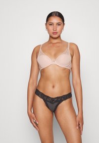 Mulher em pé de frente, vestindo um soutien nude com detalhes em renda e uma tangas cinzas escuras com renda contra um fundo simples.