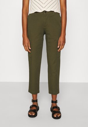 Persona con pantalones cortos verde oliva, un top de punto blanco sin mangas y sandalias negras con hebilla, de pie sobre un suelo gris.