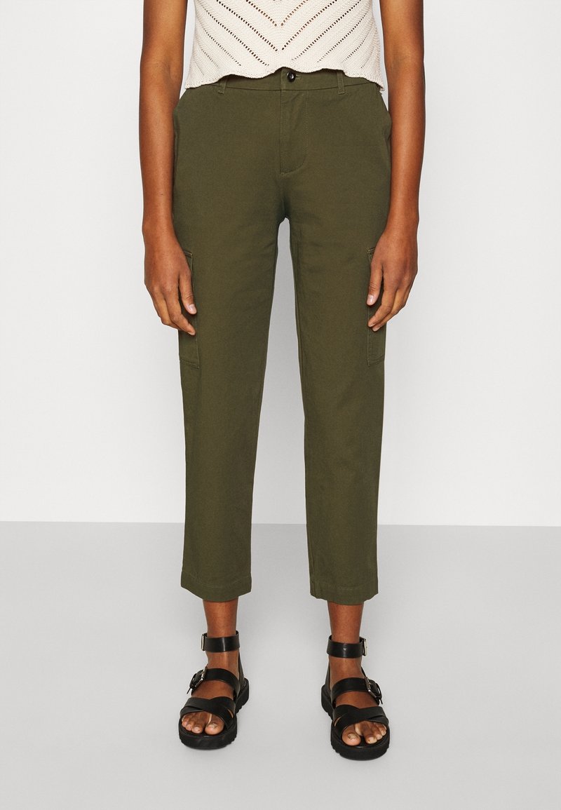 Personne portant un pantalon court vert olive, un haut blanc en maille sans manches et des sandales noires à boucle, debout sur un sol gris.