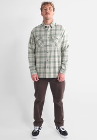 Chemise en flanelle à carreaux verts et beiges avec deux poches poitrine et un devant à boutons, assortie à un pantalon marron et des baskets noires.