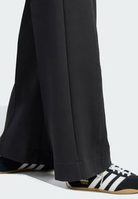 Pantalones de pierna ancha negros, hechos de un tejido suave, con un dobladillo limpio. Combinados con zapatillas negras que presentan rayas blancas y accents dorados.