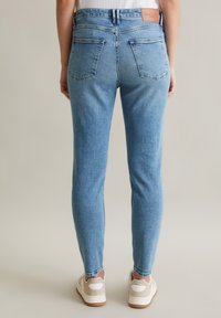 Jasnoniebieskie dżinsy o slim fit, z dwiema tylnymi kieszeniami i brązową skórzaną metką przy pasku, zestawione z beżowymi trampkami.