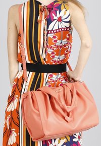 Perzikkleurige leren slouch bag met geplooide details en schouderband. Model draagt een felgekleurd jurk met strepen en bloemenpatronen.
