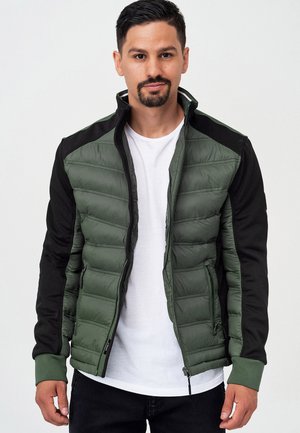 Green puffer jacket with black sleeves, zip closure, and ribbed cuffs. Features a quilted texture and side pockets. Worn over a white t-shirt.