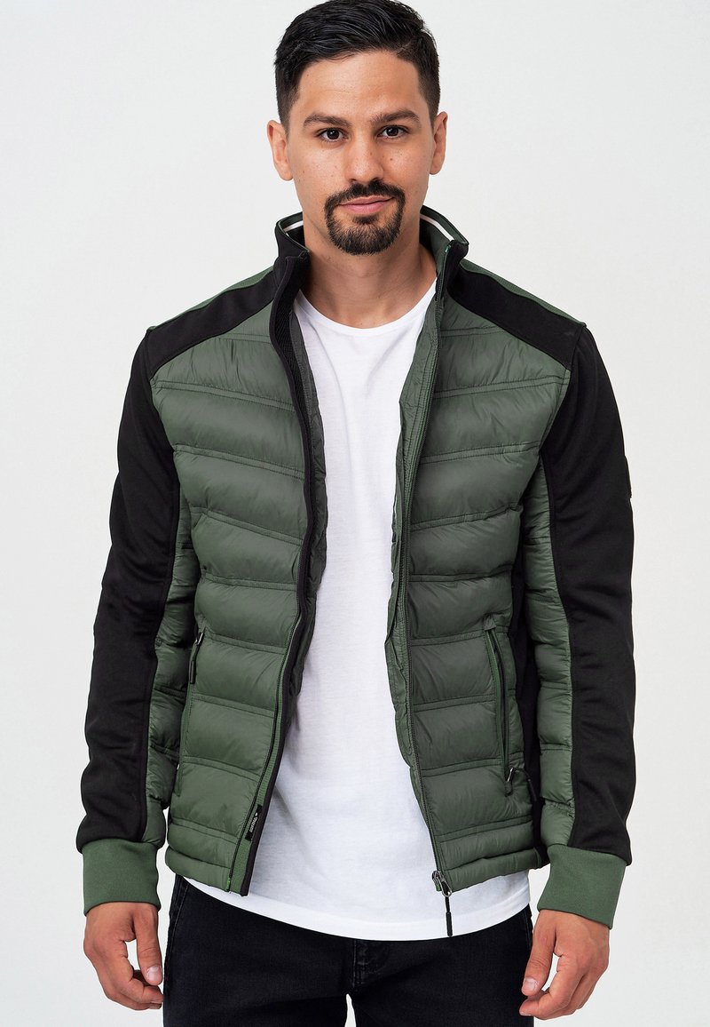 Chaqueta de plumas verde con mangas negras, cierre de cremallera y puños acanalados. Presenta una textura acolchada y bolsillos laterales. Llevada sobre una camiseta blanca.