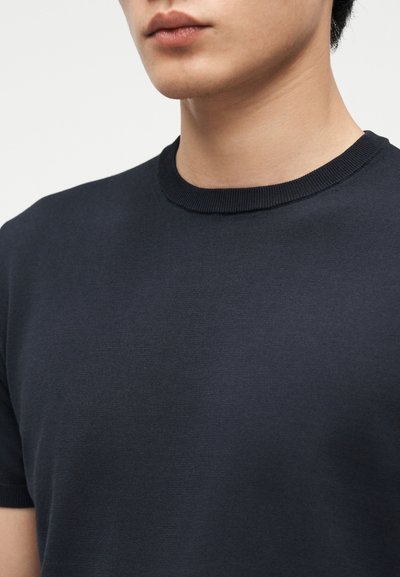 Gros plan d'une personne portant un t-shirt à manches courtes bleu marine à col rond, montrant le cou, les épaules et une partie du visage.