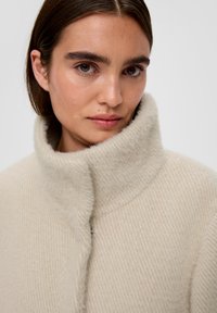 Crème kleurig fleece jack met een hoge kraag, ribbelstructuur en een zachte afwerking. Voorzien van een ritssluiting en minimale naden.