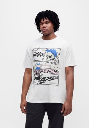Jeune homme portant un t-shirt blanc avec des graphismes de course automobile de style bande dessinée et un texte incluant "Dead serious" et "SPEEDWAY".
