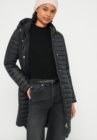 Czarna pikowana kurtka puchowa z poziomą strukturą przeszyć, kapturem i srebrnymi elementami metalowymi, noszona na czarnym prążkowanym swetrze i ciemnych dżinsach.