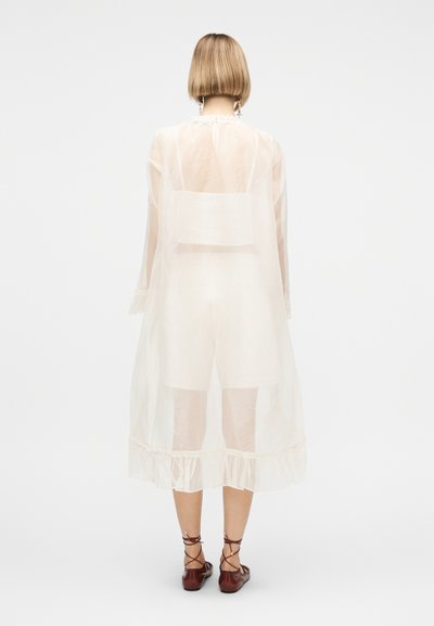 Άτομο με κοντά ξανθά μαλλιά φοράει διάφανο λευκό μακρύ παλτό πάνω από λευκά σορτς και τοπ, συνδυασμένο με καφέ δετά παπούτσια, στέκεται γυρισμένο με την πλάτη.