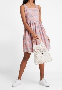 Różowa sukienka w kratkę z kwadratowym dekoltem i cienkimi ramiączkami, rozkloszowany dół. Modelka trzyma białą, dzianinową torbę. Założona z białymi trampkami.