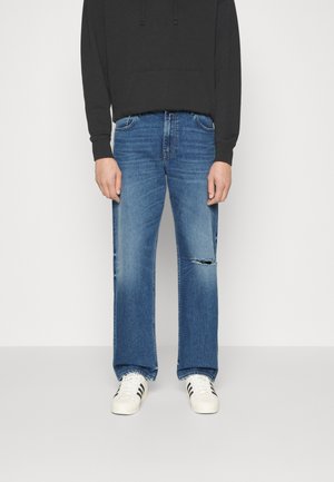 Person in ausgewaschenen blauen geraden Jeans mit einem Riss am rechten Knie, schwarzem Hoodie und weißen Sneakers mit schwarzen Streifen, stehend.