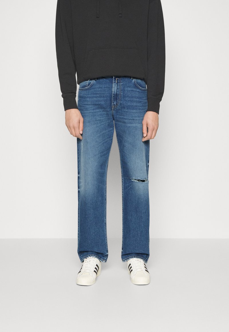 Άτομο που φοράει ξεθωριασμένα μπλε τζιν ίσιας γραμμής με ένα σκίσιμο στο δεξί γόνατο, μαύρη φόρμα και λευκά αθλητικά παπούτσια με μαύρες ρίγες, στέ standing.