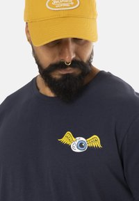 Camiseta de manga corta color navy con un gráfico de un ojo alado amarillo en el pecho. La gorra es amarilla con un logo bordado en blanco.