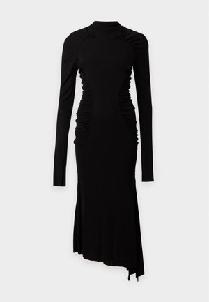 Femme aux longs cheveux foncés portant une robe midi noire moulante à manches longues avec un ourlet asymétrique et des sandales noires à talons hauts.