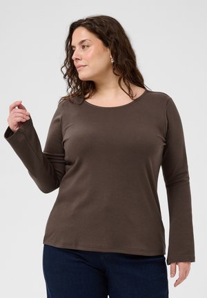 Femme aux cheveux bouclés foncés portant un haut marron à manches longues et un pantalon foncé, regardant vers sa gauche avec une main levée.