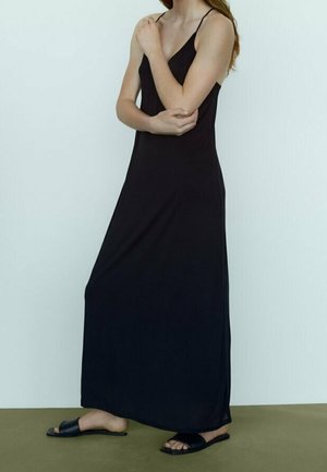 Femme en robe maxi noire sans manches et sandales plates noires, debout devant un fond blanc uni.
