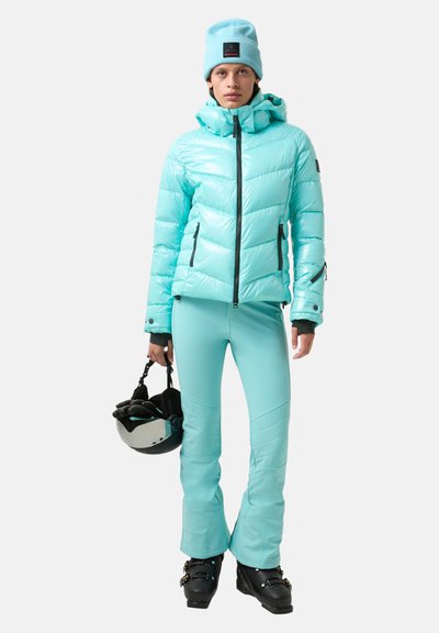 Licht turquoise donsjack met een doorgestikt patroon, gecombineerd met bijpassende nauwsluitende broek. Houdt een zwarte skihelm vast en draagt een bijpassende muts.