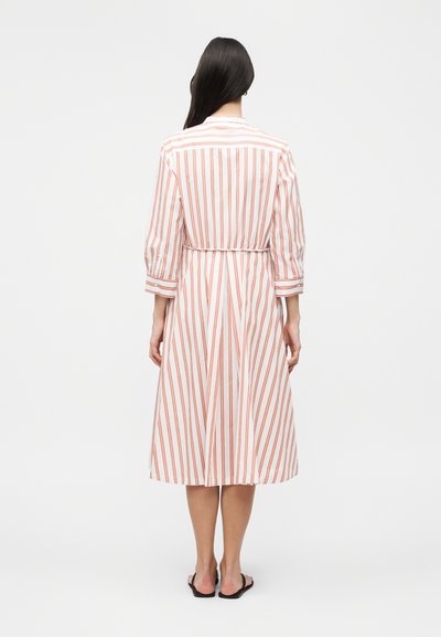 Γυναίκα με την πλάτη, φοράει ένα λευκό midi φόρεμα με κάθετες πορτοκαλί ρίγες και μανίκια τριών τετάρτων, συνδυασμένο με μαύρα φλατ σανδάλια.